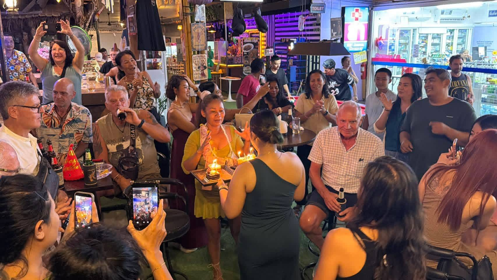 A crowd gathered around a glowing birthday cake with lit candles during a celebration at The Squirrel Bar
