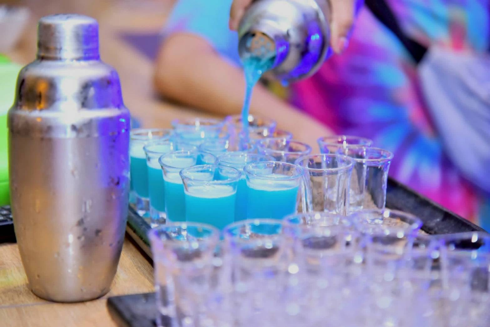 A bartender pouring vivid blue cocktails into a tray of shot glasses
