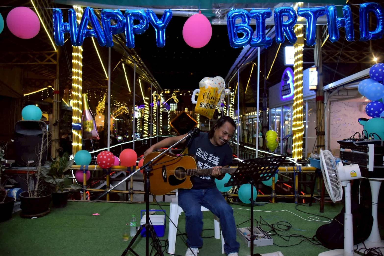 A live musician playing acoustic guitar beneath Happy Birthday balloons